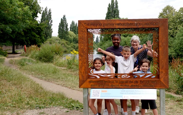 School pupils using a York rivers trail viewing frame beside the River Ouse, with riverside paths, trees and planting visible in the background.