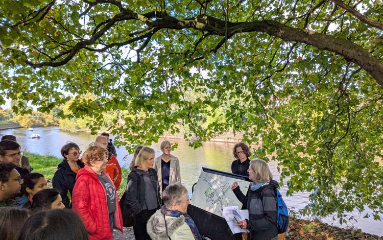 A group of people gathered under tree branches, beside the river, listening to a guide explaining a map on a display board.