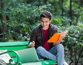 A person sits on a green bench with an orange notebook open in one hand whilst they point and smile at their laptop screen which is positioned on the bench next to them