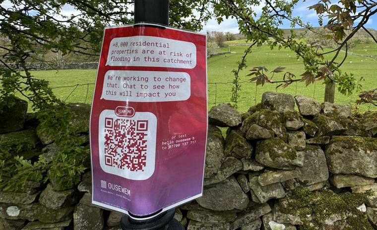 Lamp post in the countryside, with a Ousewem sign attached; the sign contains a QR code to scan and share community conversations.