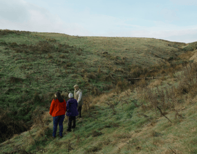 Rachel Ruth and Simon, standing on the side of a small green valley overlooking a leaky dam.